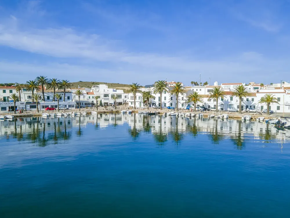 Puerto de Fornells con palmeras y casas blancas reflejadas en el agua
